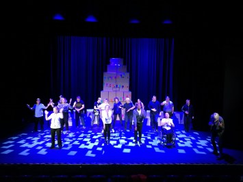 a big stage filled with people with one outstretched arm each and a monument of cardboard boxes behind them