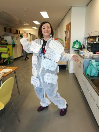 Lisette stands pen in hand with purple doc boots covered in red ladybirds. She is wearing a white boiler suit covered in giant word bubble questions such as - Who is MIMA?