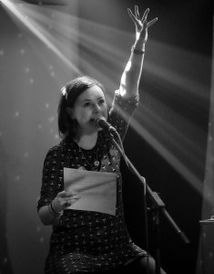 Black & white photo. Lisette sits on a stool on stage in front of a mic, spotlight on her, left arm reaching up.