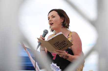 Lisette with mic and book saying 'Stay Focused + Extra Sparkly' pulling bizarre faces on a festival stage with comic book dress and gold hologram backpack.