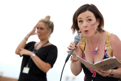 Lisette with mic and book saying 'Stay Focused + Extra Sparkly' pulling bizarre faces on a festival stage with comic book dress and gold hologram backpack.