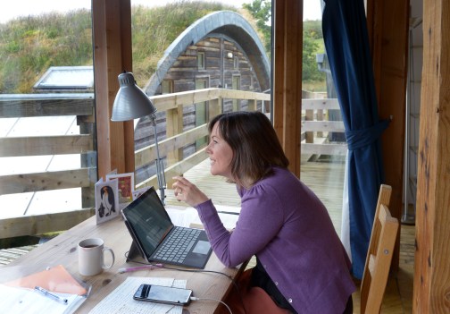 photo description: I'm sitting at my desk in my studio, wearing a purple cardigan. The desk has a lamp, laptop and phone, a mug with a lipstick mark and lots of parers and pens on it. In the background you can see through the window to an adjacent hobbit hut with grass growing on its sloped roof. photo credit: Alan Dimmick