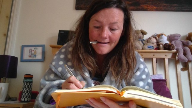 photo description: Lisette's hair is messy. She is wearing her dressing gown and is working in bed. Soft toys on the headboard behind her, a bedside table with penguin covered water bottle, an alarm clock and a lamp. She has a pen top in her mouth, she is holding her pen and concentrating on writing in a yellow notebook.