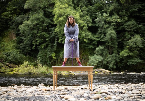 photo description: Lisette, a white woman with freckles and long brown hair, stands on top of a wooden table. She is wearing a grey dressing gown, purple Dr Marten boots and leaning on a purple perspex walking stick. The table is on a pebble beach with a river running behind it, with lots of green trees. image credit: Rob Irish