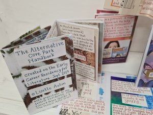 photo description: 6 handmade multicoloured little instant books, most blurry, to the Foreground The Alternative Cove Park Handbook which is blue handwriting on a background of photocopied leaves