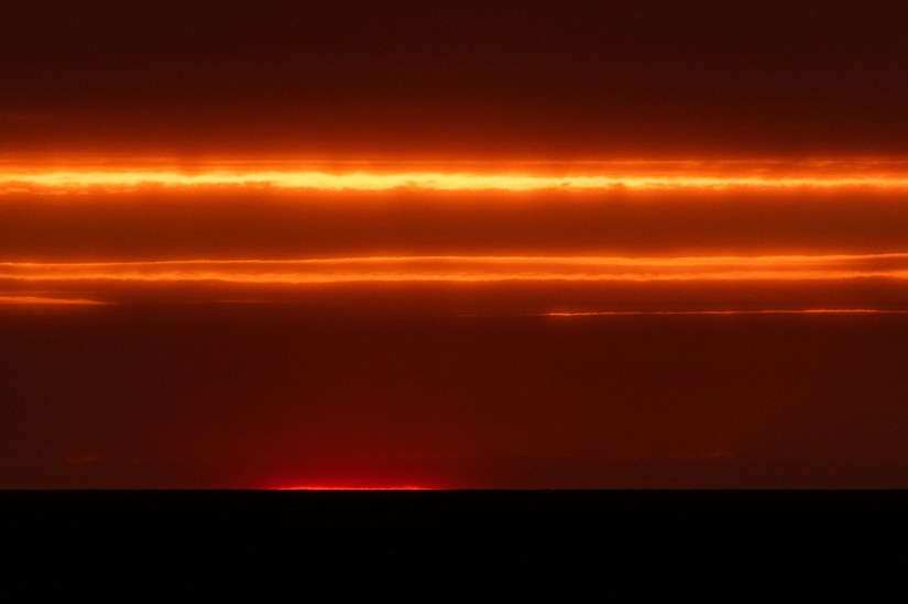 photo description: landscape. Black horizon. Sky striped in fiery reds and oranges. Sun just to say creeping up. Photo credit: Kev Howard