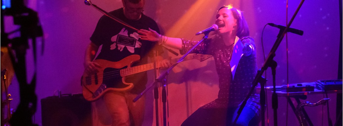 Lisette sitting behind a microphone, shouting, arms outstretched. John Pope on bass guitar behind. Stage lit in pinks and purples.