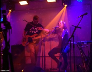 Lisette sitting behind a microphone, shouting, arms outstretched. John Pope on bass guitar behind. Stage lit in pinks and purples.