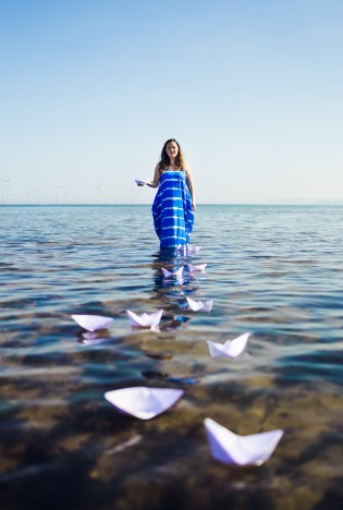 photo description: Lisette wears a sea blue dress with whit horse waves, stands in the shallows holding a paperboat with more floating in front of her. Wind turbines stand behind. photo credit: Rob Irish